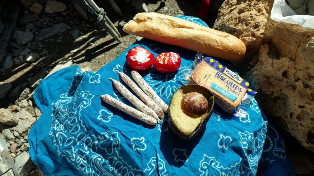 Blue bandana topped with picnic lunch of baguette, sausages, crackers, cheese and avocado.