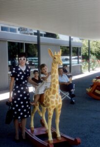 Woman in cateye sunglasses and black poka dot belted dress, black high heels, standing next to small child who is sitting on a toy giraffe.