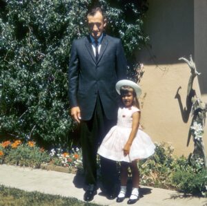 Man in dark suit with young daughter dressed up in Easter best with a pink dress, white hat and black patent leather shoes with white socks.