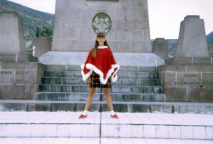 A girl of 10 wearing a bright red poncho trimmed with white fake fur stands on the equator in Equador