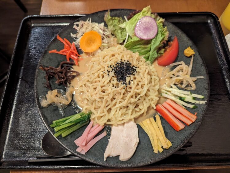 Plate of cold noodles surrounded by groups of colorful vegetables around the side: tomato, carrot, cucumber, pickled vegetables