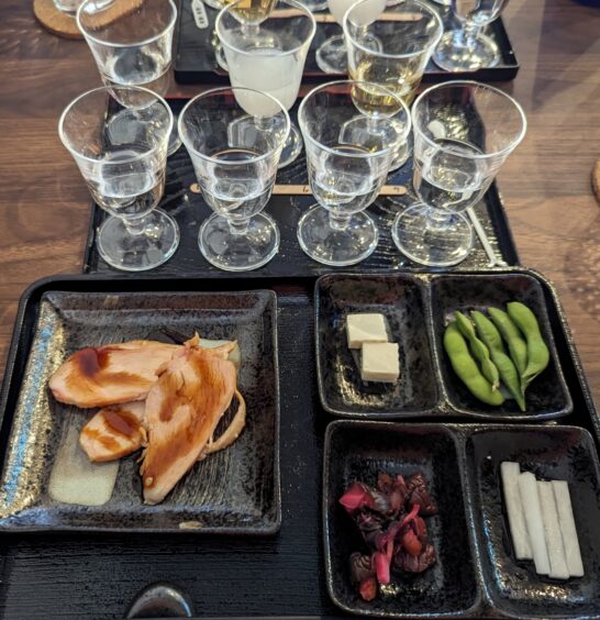 Seven glasses with different sakes with five dishes of food: seafood, tofu, pickled vegetable and edamame