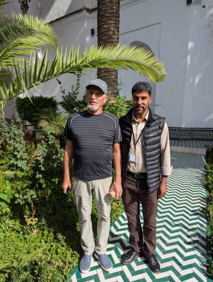 Two men standing in a garden under a palm tree. One is wearing a guide badge. 