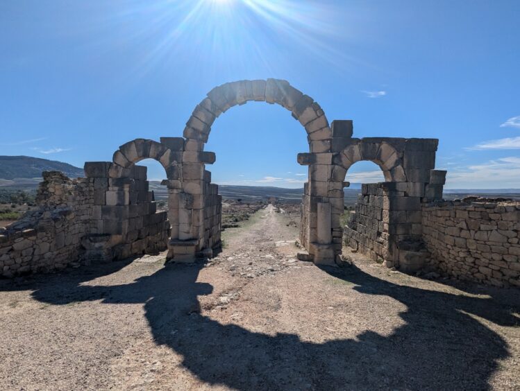 Roman arch ruins framing the remains of a village with fields in the background