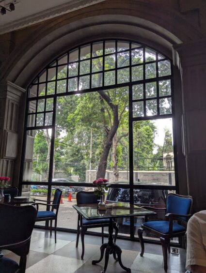 Table and two chairs in front of a large window looking out into a leafy street. 