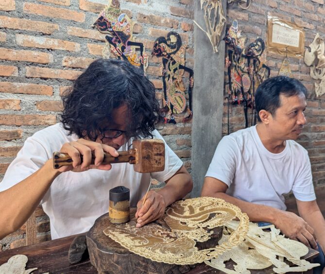 A male artist bends over a piece of leather as he uses a wooden mallet and sharp object to create the traditional design for a shadow puppet.