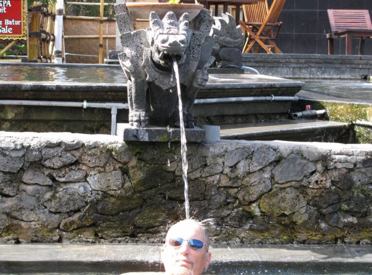 Man in pool of water sitting under a spout of water from a fountain in the shape of a mythical animal