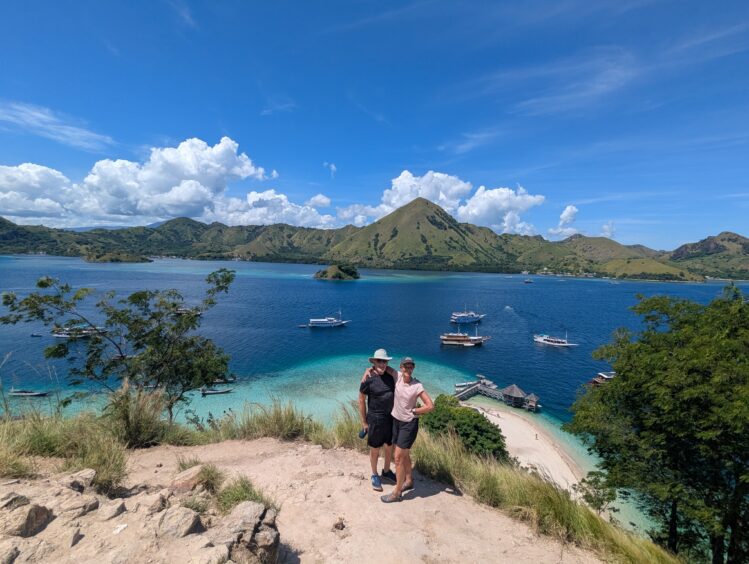 Man and woman on a hill with a panoramic view of turquoise and blue water and green hills
