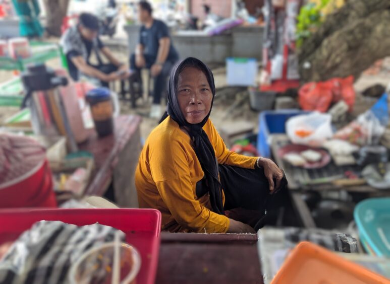 Woman with black scarf and orange shirt sits at her stall surrounded by foods for sale