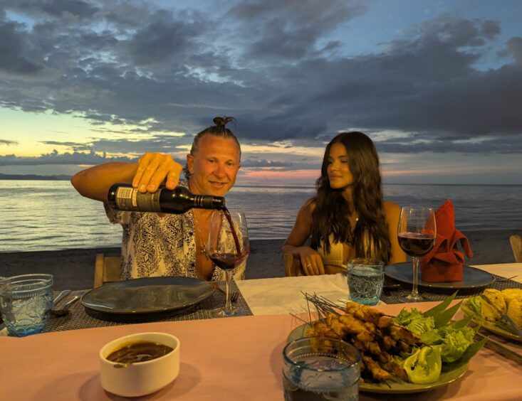 A man pours wine for a woman at a table on the beach with the water and sunset behind them, sate skewers in front