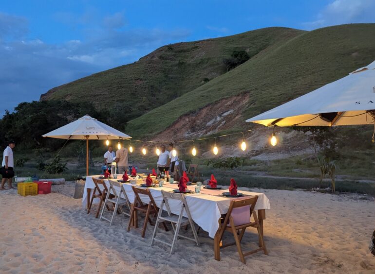 A table decked out with white tablecloth, red napkins and a string of lights suspended above on a beach