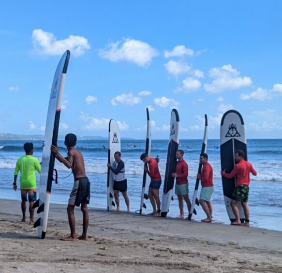 Five surf students and two instructors lined up on the beach holding their surfboard upright